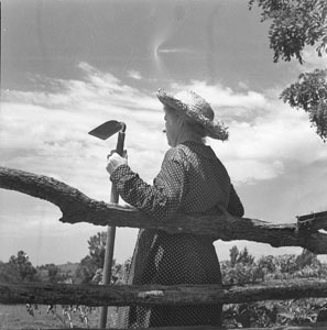 Woman Hoe in hand back to camera leaning against rail-fence